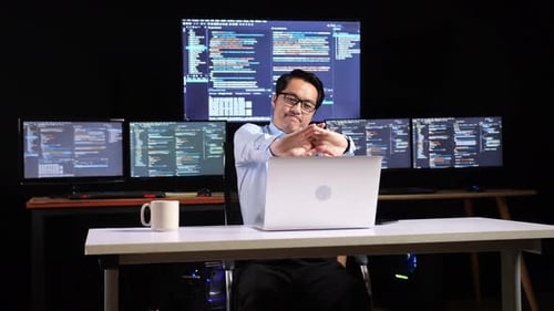 Asian Male Programmer Stretching While Writing Code By A Laptop Using Multiple Monitors