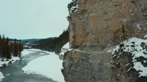 Drone shot rising up the side of a snowy cliff face with a frigid creek below with Canadian goose in