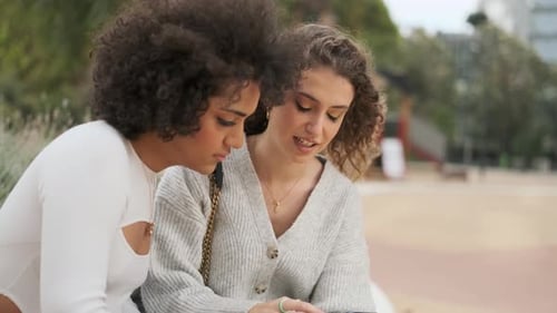 Two Friends Using a Mobile While Sitting on a Bench in a Park
