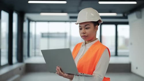 Confident Female Engineer in Orange Vest Inspects New Office Building with Laptop and Friendly Smile