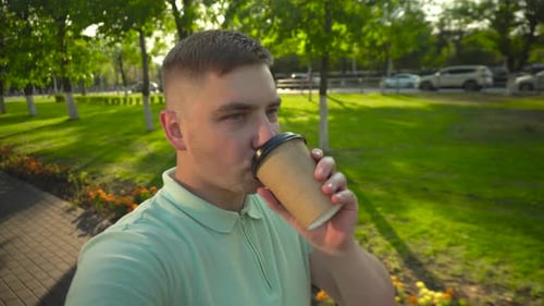 A Young Man Walks Along a Green Alley with Coffee in His Hands A Man Drinks Coffee and Films a Vlog