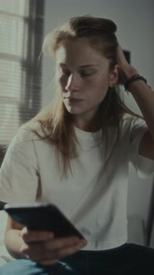 Pensive Woman Sits With Phone in Bedroom