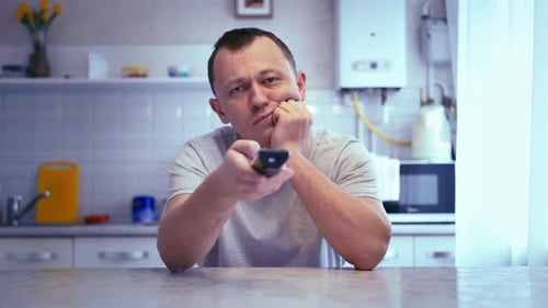 Homem melancólico sentado na cozinha de casa, distraidamente, navegando no canal, perdido em pensamentos