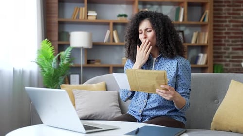 Disappointed young woman reading letter with bad news sitting on sofa at home office. Upset female