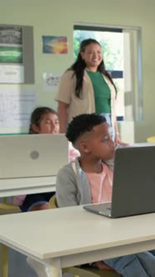Vertical video: Smiling teacher in green top guiding boy looking at laptop in class with tablet