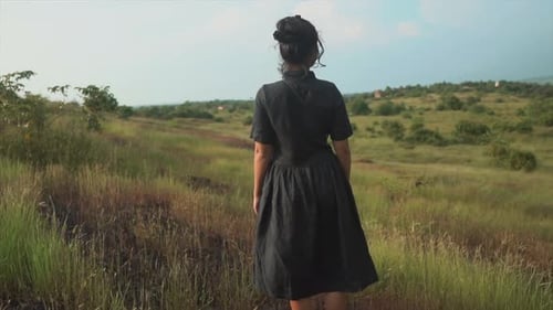 Brunette woman in black dress standing on a green meadow and blue sky background.