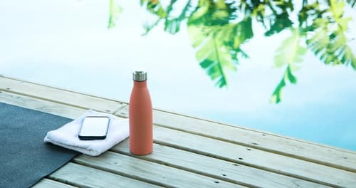 Yoga mat, water bottle, and smartphone on deck by pool, tropical setting, copy space
