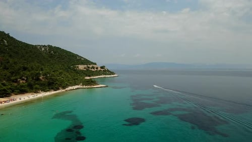 A serene beach with turquoise waters, green hills, and a boat leaving trails under a clear sky.