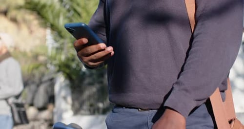 Hombre afroamericano agarrando una bicicleta desplazando textos en el teléfono, sonriendo y usando una bolsa en la acera