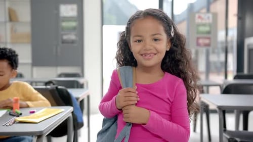 In school classroom, young biracial girl holding a backpack smiles