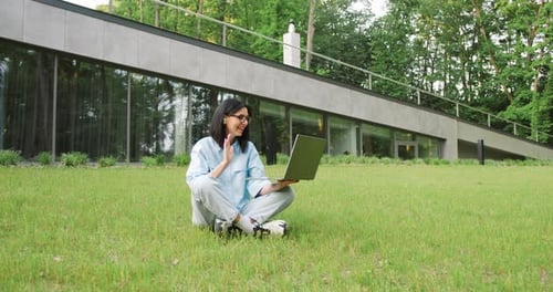 Young female freelancer working remotely in a city park, sitting on the grass and engaged in a video