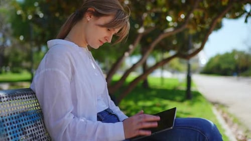 Professional Woman Working on Tablet Sitting on Sunlit Park Bench Relaxing Amid Green Surroundings