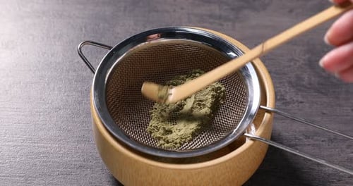 Woman sifting green matcha powder into ceramic bowl at gray textured table, closeup