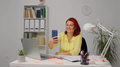 Smiling young red-haired woman making video call while sitting at desk in home office.