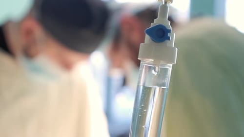 Close Up of Intravenous Drip Chamber with Medicine Drops and Surgeons Working in Background in