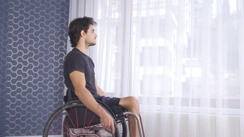 Young disabled person sitting in a wheelchair watching out the window at home.