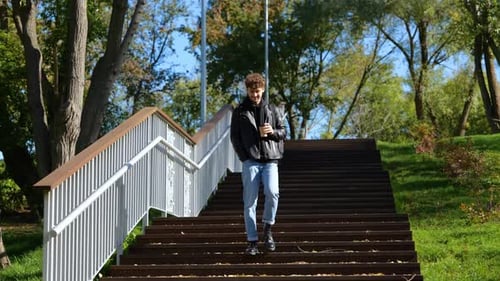 Happy Young Man with Backpack Going Down Stairs and Holding Coffee in the Park Enjoying the View