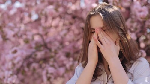 Irritated Caucasian Young Woman Standing Near Blooming Sakura Tree and Scratching Itchy Eyes During