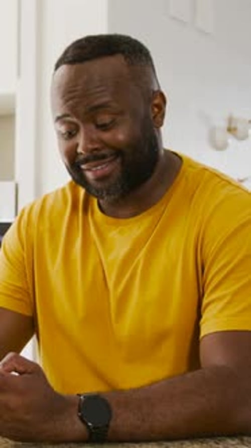 Vertical video: Picking up device, man in yellow tee leaning on counter, checking smartwatch screen