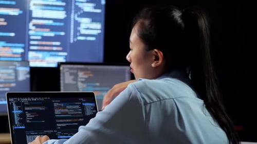 Vue arrière rapprochée d'une programmeuse asiatique appuyée sur une table tout en écrivant du code par un ordinateur portable