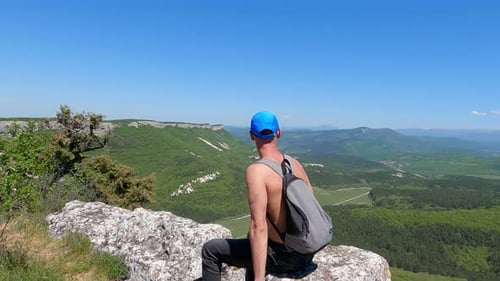 Shirtless Tourist with Backpack Sitting on Rock While Looking at Green Valley