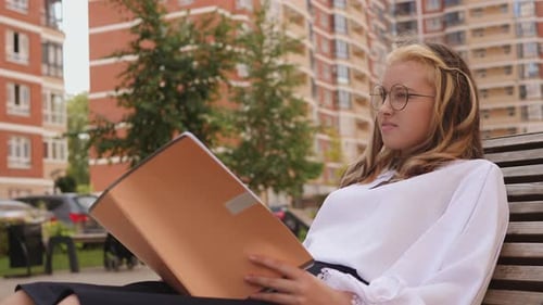 A Girl in a School Uniform is Sitting on a Bench with a Notebook and Reading