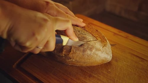 Close-up hands of female baker with knife cutting loaf of bread to slices at bakery or kitchen
