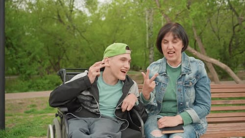 White Caregiver And Young White Man In Wheelchair Sharing Cheerful Chat On Park Bench Playful Faces