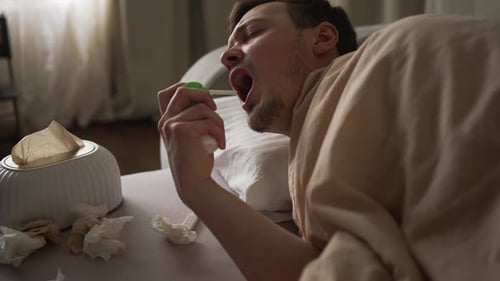Un homme souffrant de symptômes du rhume applique un spray pour la gorge alors qu'il est allongé dans son lit luttant contre une infection virale et