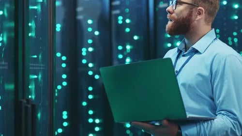 A Focused Bearded Man Works in a Server Room with a Laptop