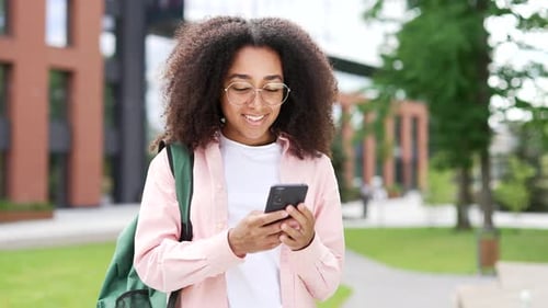 African american female student using phone standing on campus space near university building. Happy