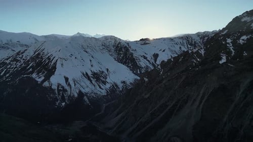Aerial snowy winter mountain scenery with steep valleys, sun dissapearing behind ridgeline