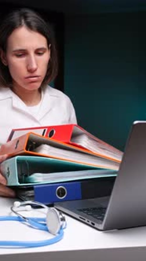 Vidéo verticale : médecin regardant une pile de dossiers médicaux sur un bureau avec un ordinateur portable