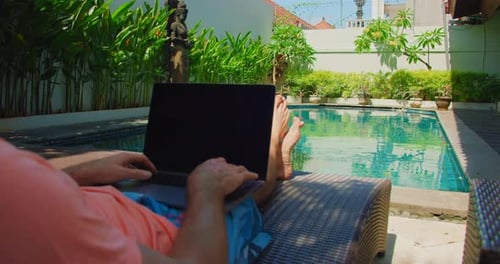 Man Freelancer Sits on Deck Chair Near Swimming Pool and Works on Laptop