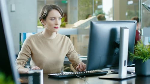 Schöne junge kaukasische Frau sitzt an ihrem Schreibtisch und arbeitet an einem PC. Im Hintergrund