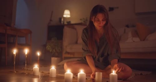 Happy Brunette Girl in a Green Shirt Sits on a Blanket on the Floor and Lights Candles to Create a
