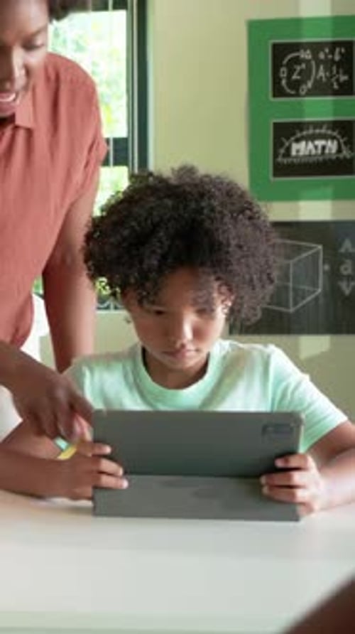 Vertical video: Teacher entering left, student in mint tee adjusting tablet in class for math
