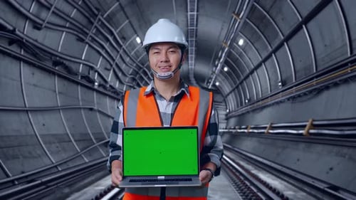 Asian Male Engineer Smiling And Showing Green Screen Laptop To The Camera In Subway Tunnel