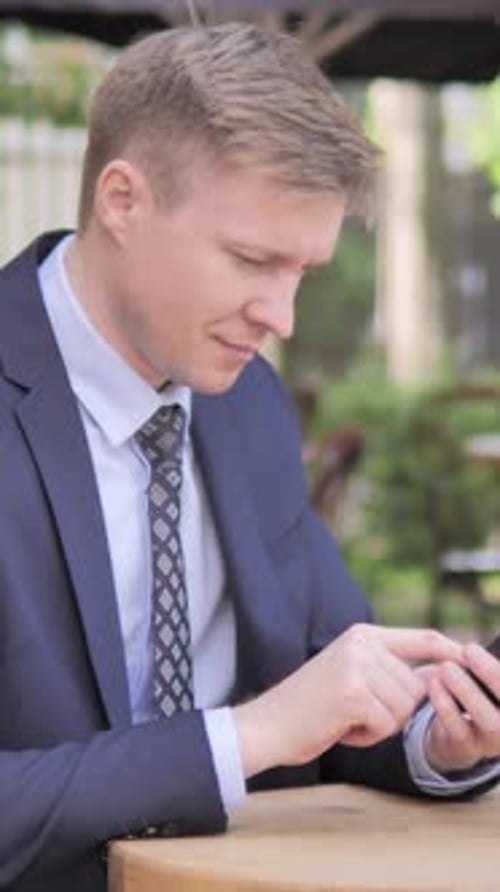 Businessman Upset by Loss on Smartphone, Sitting in Outdoor Cafe, Vertical Video