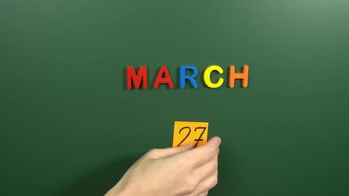 Hand sticking a sticker 27 March calendar day on school board. 27 date of March. Twenty seventh da
