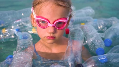 A little girl swims among plastic garbage in the ocean.