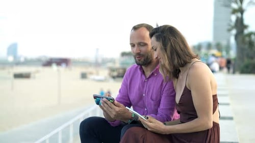 Young couple relaxing with smartphone on summer beach in coastal city