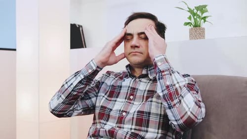Man Sitting on a Couch Holding His Head in Discomfort While Experiencing Stress in a Modern Indoor