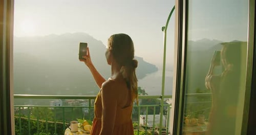 A woman in a yellow dress takes a photo with her phone from a balcony overlooking the Amalfi coast a