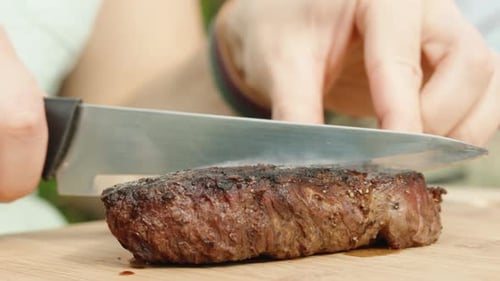 I cut a piece of beef steak in half with a knife on a wooden cutting board, close-up