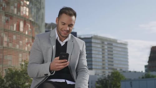 Smiling Businessman Typing on Smartphone at Street. Confident Executive Sitting on Bench