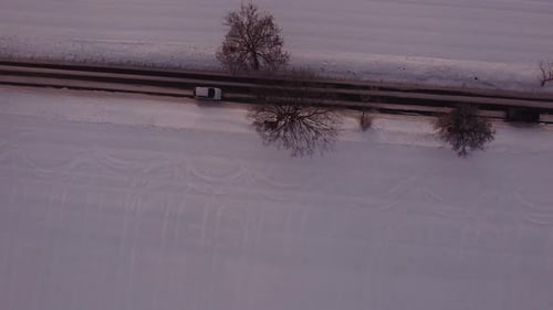 Cars drive along not fully cleared of snow local road between snow-covered fields. Aerial top down v