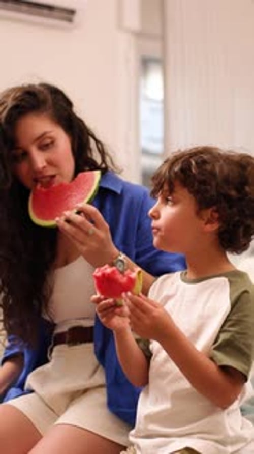 Woman and boy sit eating watermelon in home