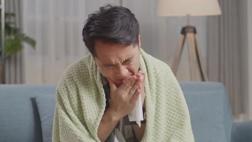 Asian Man Suffering From Cold On Sofa At Home. Sick Guy Blows Sneezes Wipes Snot Into Tissue