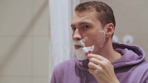 Homem se barbeando no banheiro com espuma no rosto e expressão relaxada enquanto se prepara para o dia